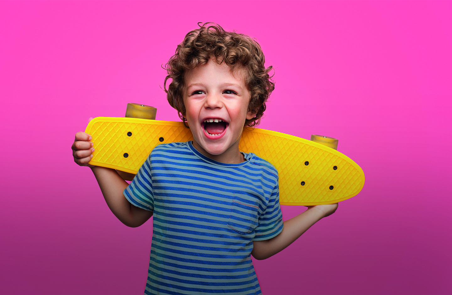 Ein Junge hält mit beiden Händen ein gelbes Skateboard hinter seinen Rücken, vor einem pinken Hintergrund