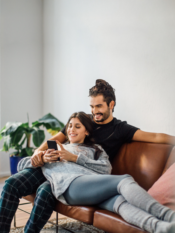 ein Mann sitzt auf der Couch, eine Frau liegt auf seinem Schoß und schaut in ihr Handy