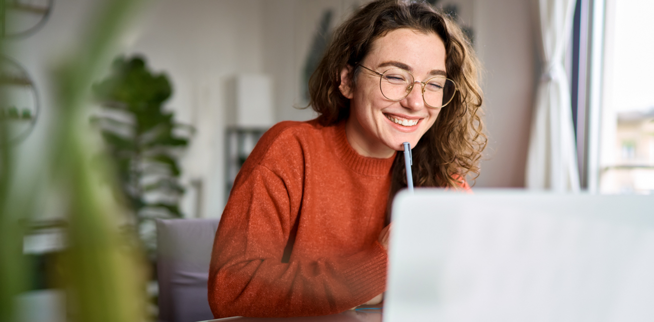 Eine junge Frau im orangenen Pullover sitzt lachend vor einem Laptop.