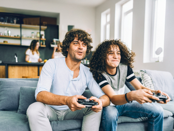Ein Vater und sein Sohn sitzen auf einer Couch und spielen Videospiele mit Controller in der Hand.